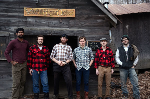 Six men in plaid shirts stand in front of a sugar shack. A sign behind them reads "West Hill Sugar Orchard".